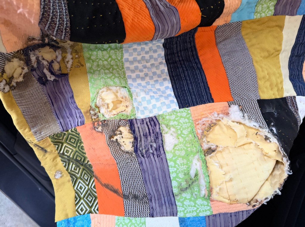 Close-up photograph of a vintage handmade quilt with visible burn damage and torn fabric patches, showing exposed batting and worn stitching from a past house fire. The quilt rests in natural light, reflecting age, memory, loss, and the possibility of restoration.
